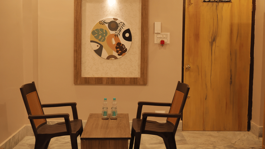 2 wooden chairs facing each other with a small table between them, with a king size bed, and a wall art, in a room at Shophori Wellness Retreat, Bolpur.