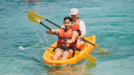Two people kayaking in Vietnam, in clear blue water, with other kayakers and swimmers visible in the background.