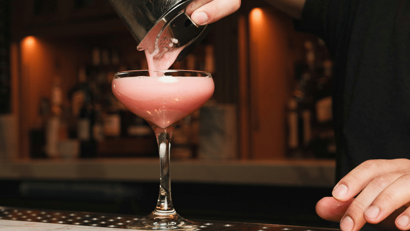 High-contrast close-up shot of a bartender pouring a pink cocktail into a coupe glass.