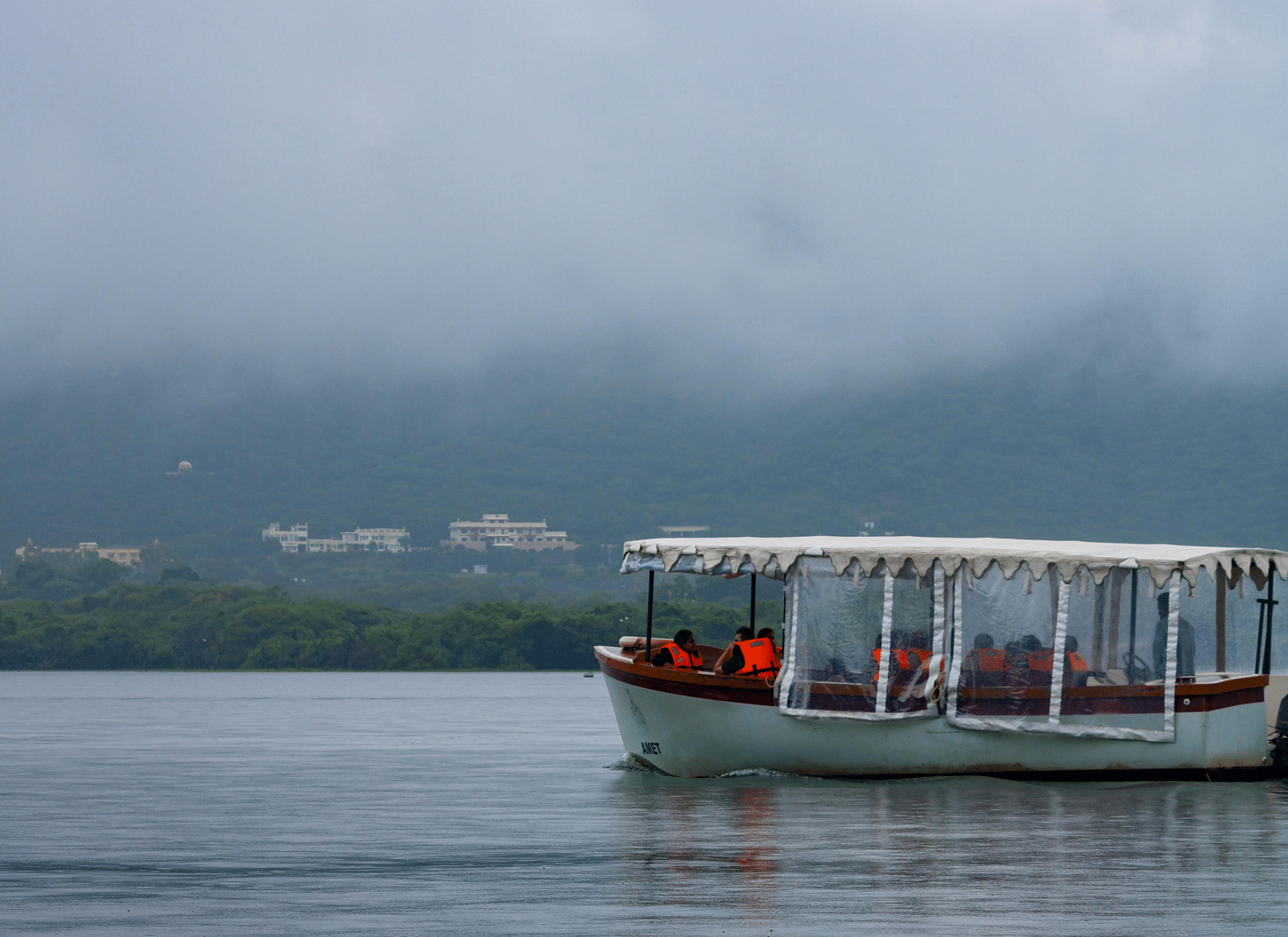 A small, enclosed tour boat with visible passengers, some wearing orange, navigates a calm lake under a cloudy, overcast sky, with a hint of distant land barely visible on the horizon.