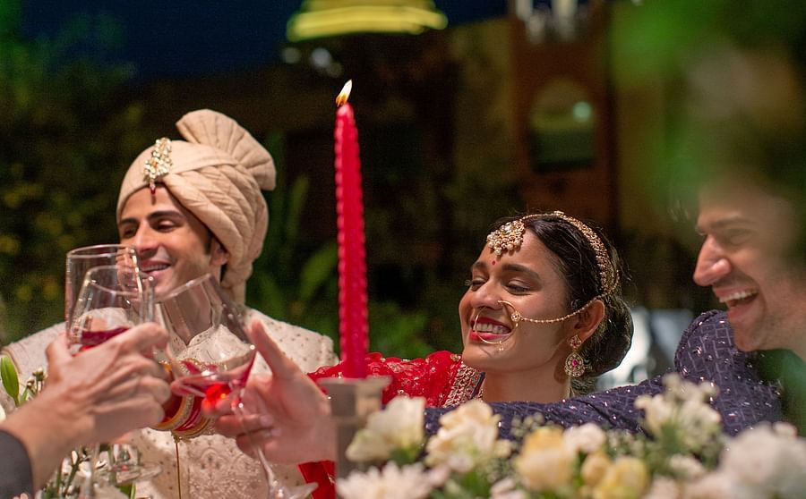 A joyful wedding scene with a bride and groom, the groom wearing a turban, both smiling and participating in a toast under a floral arrangement and string lights at night.