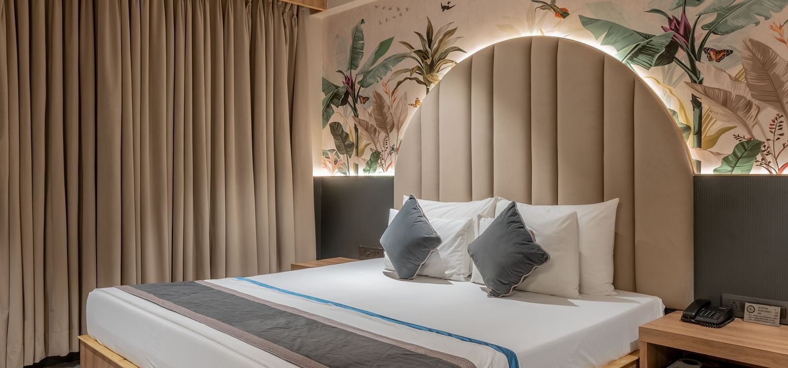 A bedroom with king size bed, ceiling fan, a wooden side table, and curtain spread across the window at Hotel Highway King, Jaipur.
