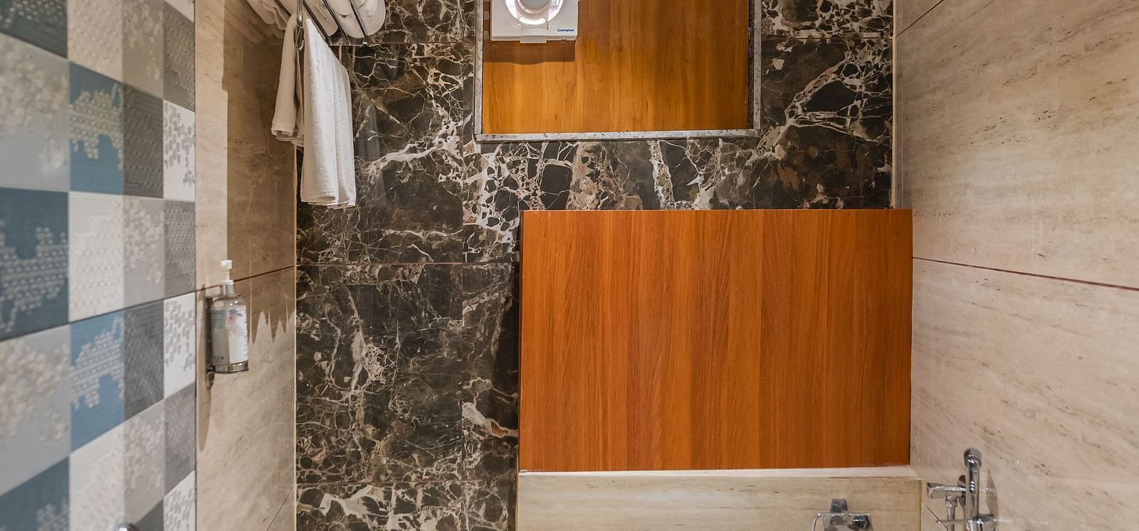 A compact bathroom features a shower area with patterned tiles, a toilet, and a wooden-paneled section - Lords Eco Inn Ambaji.