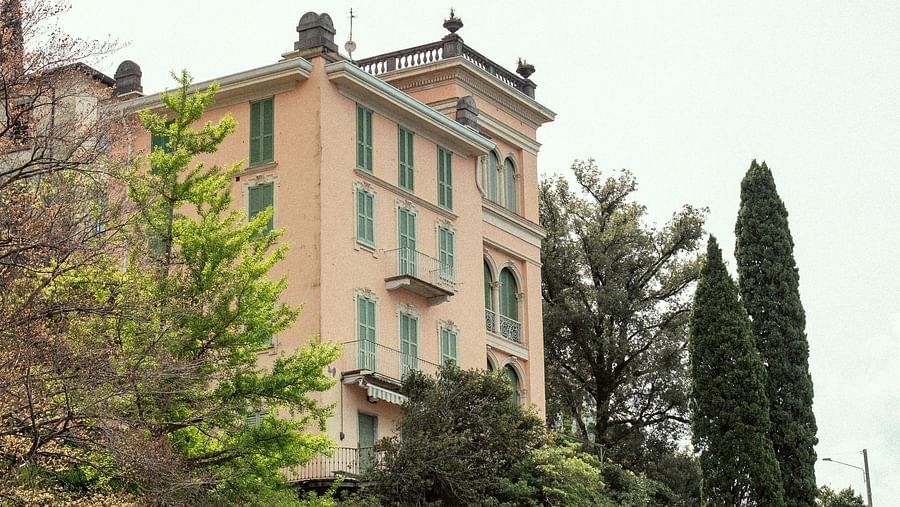 A stately, light-colored building with multiple stories and green shutters stands on a hill surrounded by lush trees under a pale sky.
