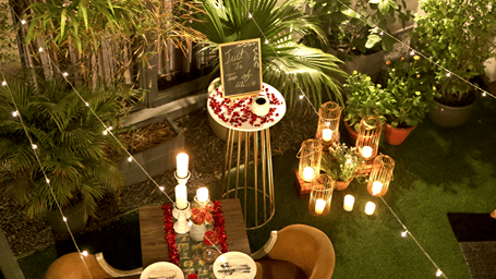 An intimate outdoor dining setup with a table for two, candles, floral petals, decorative lanterns, string lights, and potted plants.