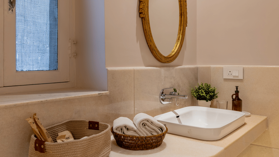 A bathroom wash counter at Barwara Kothi, Jaipur featuring a square basin, ornamental mirror, towels in baskets, and side window.