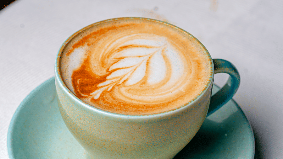 A cup of latte with leaf-patterned foam art at Café Latte, located within Noor-Us-Sabah Palace, Bhopal, served on a light green saucer.