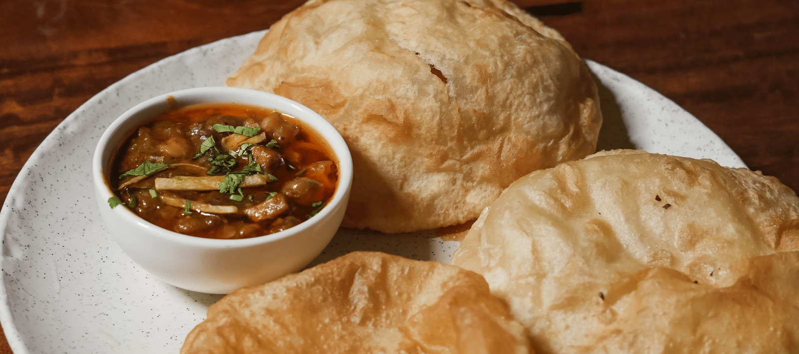A dish of two golden-brown, puffed bhature breads served with a small bowl of chickpea curry (chole) on a white plate at BluSalzz Select City Centre, Amritsar.