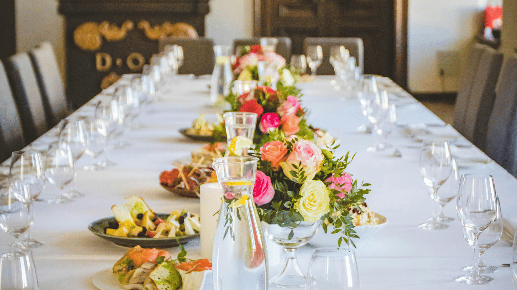A long dining table decorated with flowers in the middle along with shiny glassware and cutlery set on the table.