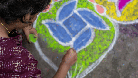 A woman kneels on the ground, filling a large, colourful kolam design with bright powders.