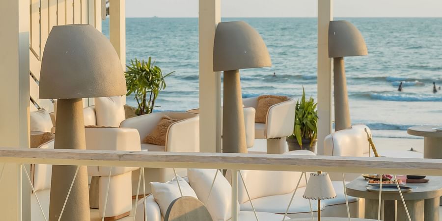 An elegant outdoor dining or lounge area at Living Room Hotels and Resorts featuring white tables and chairs, under a canopy, overlooking the ocean and palm trees.
