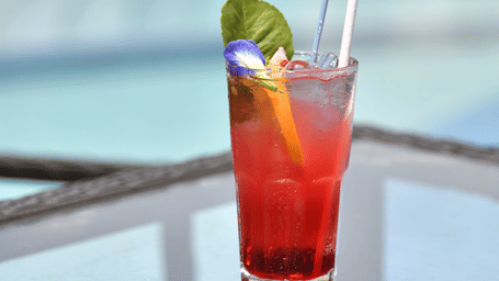 A tall glass of red drink garnished with a green leaf and straw placed beside the swimming pool at Vedic Village Spa Resort with a blurred poolside background.