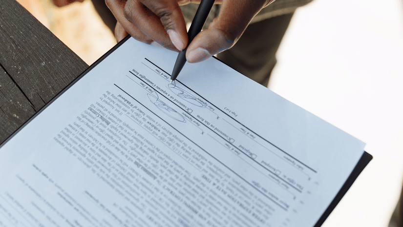 A person uses a black pen to sign a paper document on a clipboard.