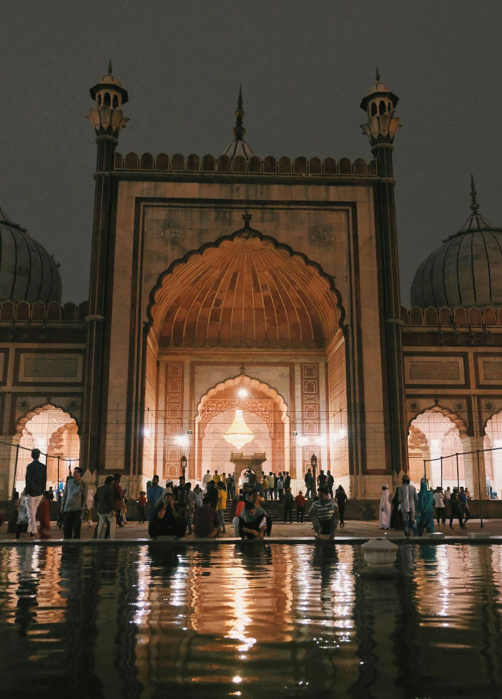 A masjid view with lights on and a grey sky background and packed with pilgrims and some can be seen washing their hands at the water body in front of the masjid.
