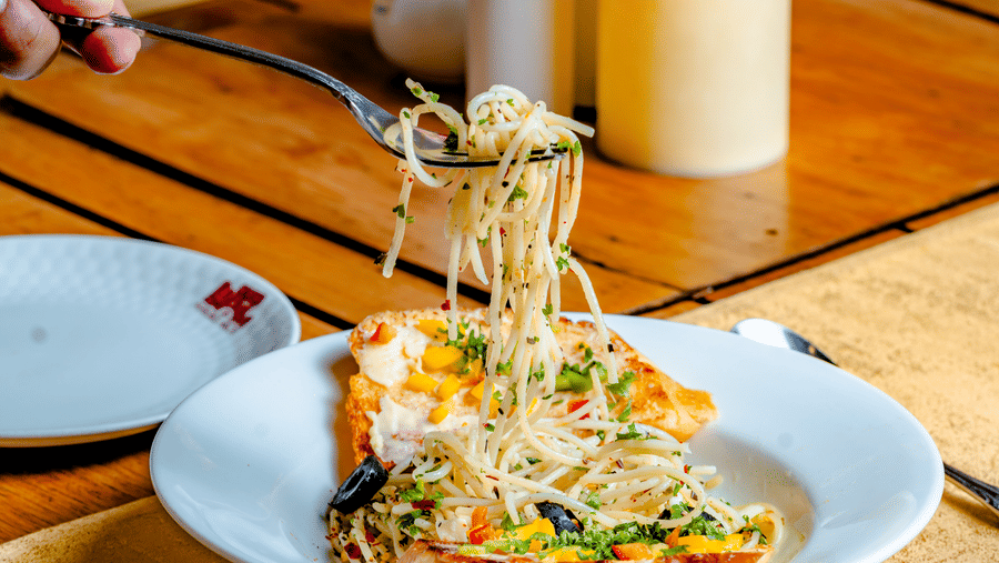 A person lifting pasta with a fork at Marble Arch, located within Noor-Us-Sabah Palace, Bhopal, served on a laid table with flowers and a candle.