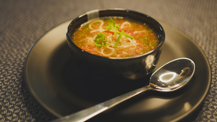 A bowl of hot, delicious soup served with a spoon on a dark textured plate under warm lighting.