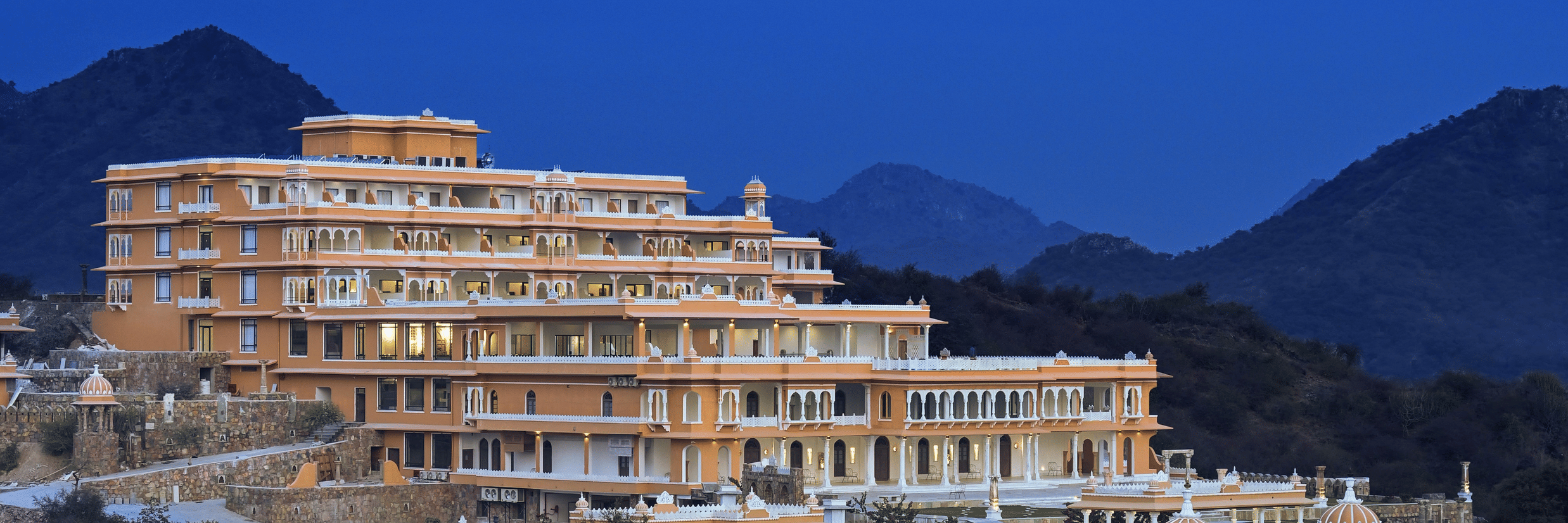 Facade of Fateh Vilas during twilight with numerous trees in the foreground and mountains in the background, ideal for a destination wedding in Udaipur