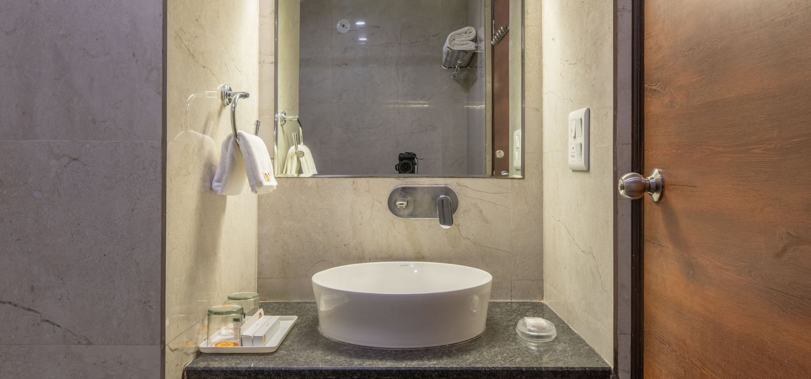 A narrow bathroom with washbasin, mirror, and wooden door at Hotel Highway King, Jaipur.
