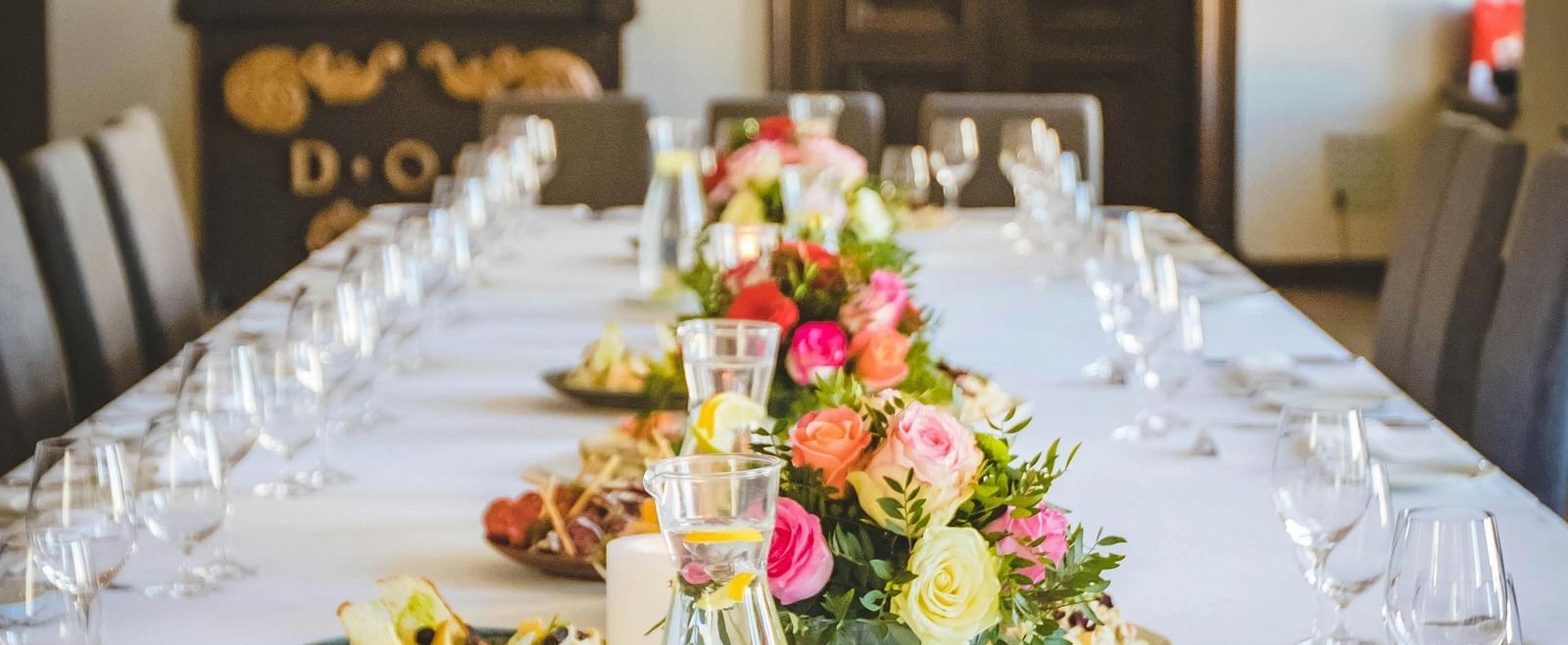 A long dining table decorated with flowers in the middle along with shiny glassware and cutlery set on the table.