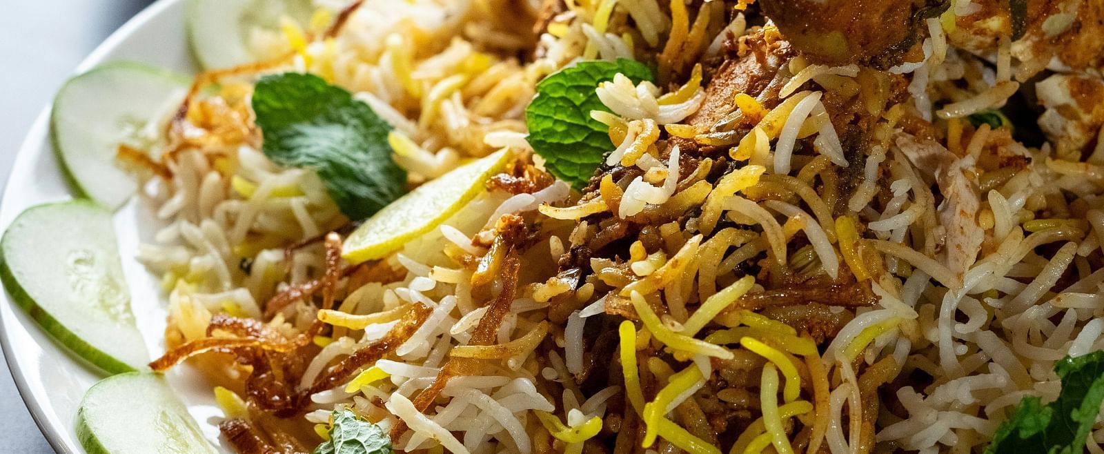 A plate of chicken biryani garnished with fresh mint leaves, fried onions, and cucumber slices, served with bowls of raita and curry on the side.
