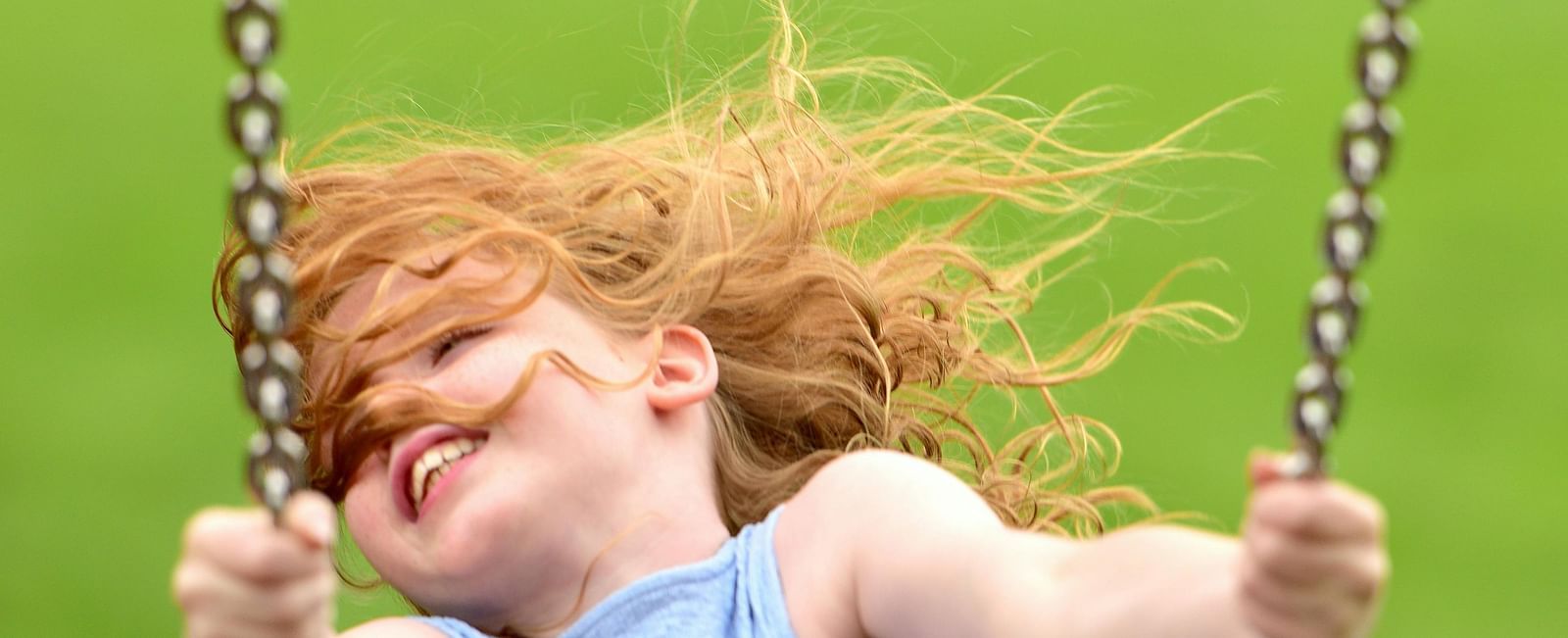 A young girl with red hair, wearing a light blue dress, is captured mid-swing on a park swing, against a blurred green background.