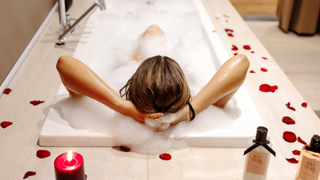 A person reclining in a rectangular bathtub with their arms behind their head at SUKAVI, surrounded by flower petals arranged around the bath area.