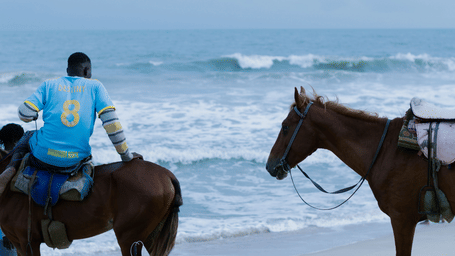 A man sitting on a horse at the beach, in front of the water as another horse stands on the side.