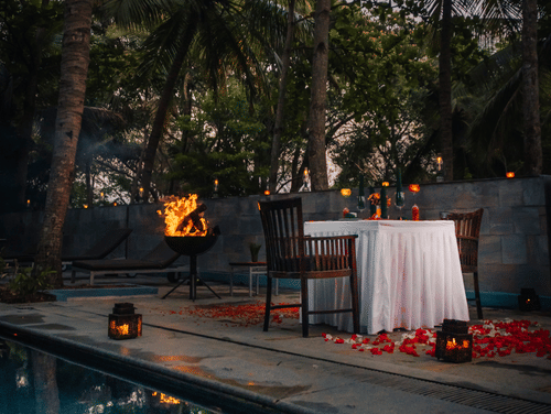 A romantic, private dinner set up poolside at dusk, surrounded by lush tropical greenery and tall palm trees.