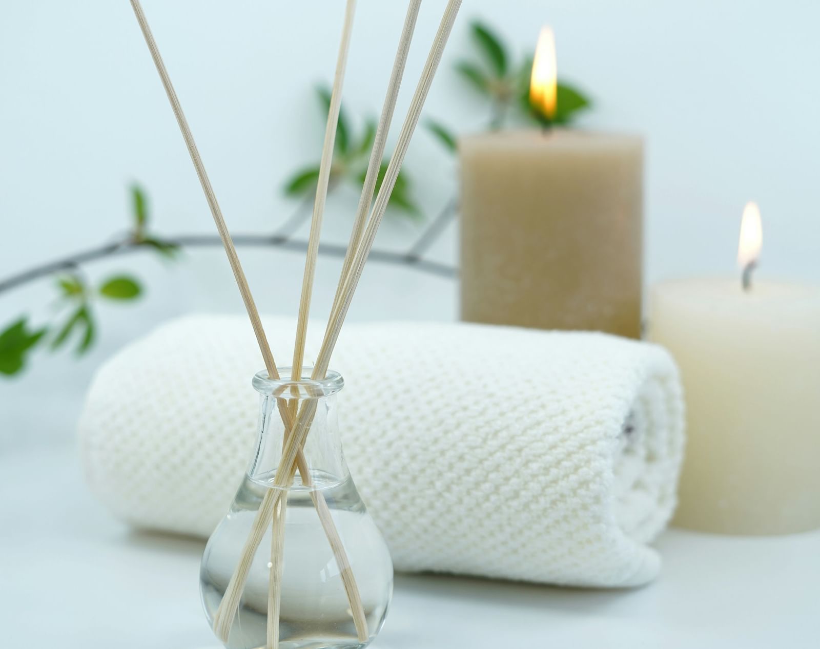 An arrangement of spa or wellness items, including a reed diffuser, a lit candle, and rolled white towels, set against a light background.