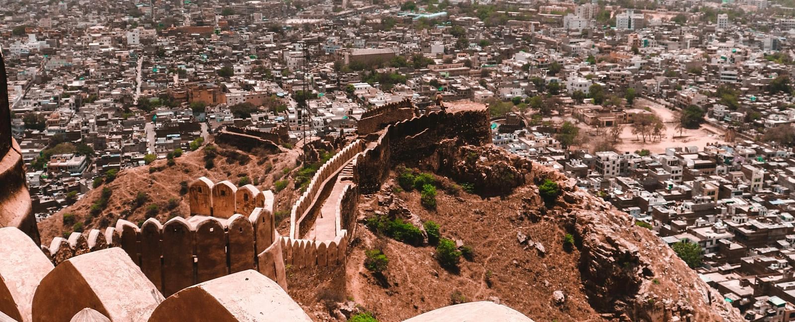A high-angle view from Nahargarh Fort, Jaipur, overlooking the sprawling city with its densely packed buildings and a distant mountain range under a cloudy sky.