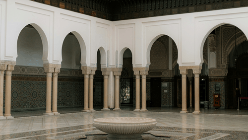A brightly lit courtyard with a fountain in the center, surrounded by vaulted arches and a tiled floor.