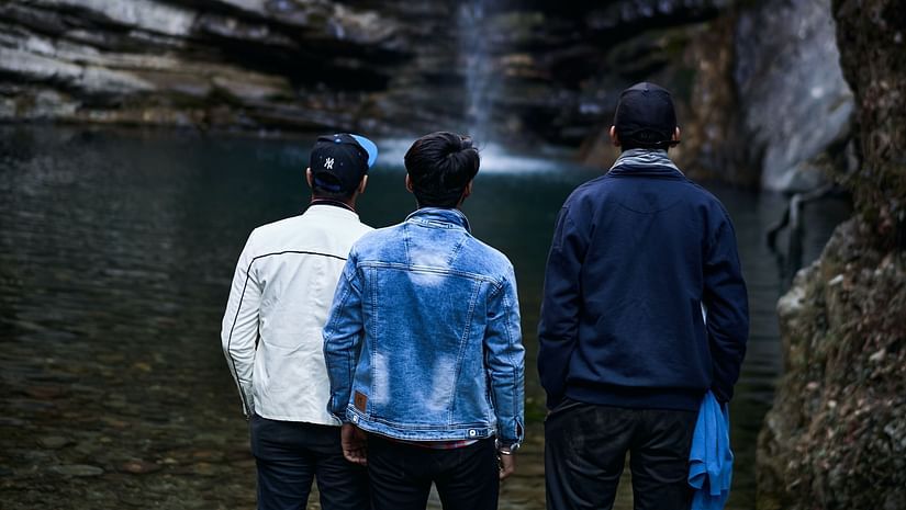 Three individuals stand on dark rocks, gazing up at a cascading waterfall that plunges into a shadowy area below. They appear to be enjoying the natural beauty of the scene