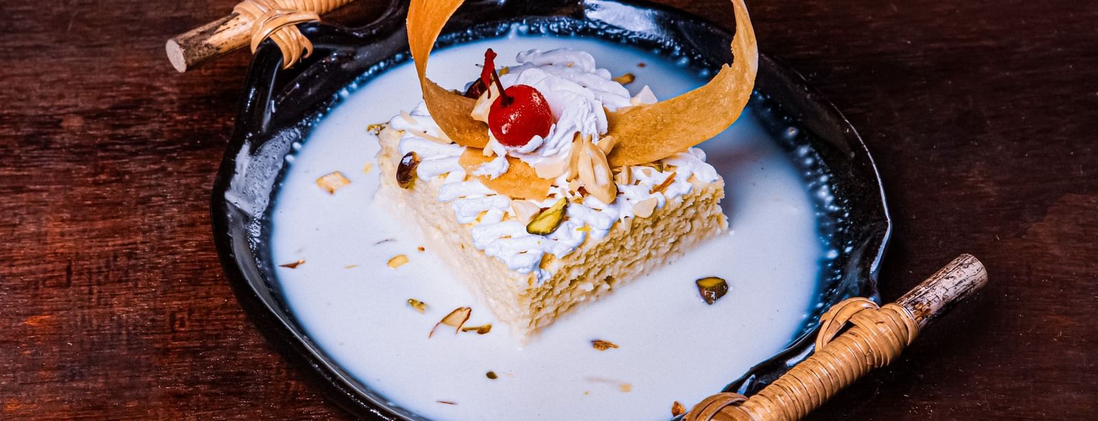 A plated dessert with a decorative basket element, garnished with cream and a cherry, presented on a white plate with sauce swirls on a dark wooden surface at The Hamuse Hotel, Kodaikanal.
