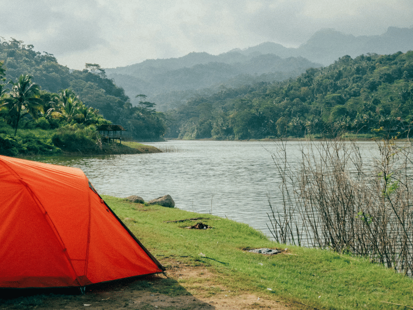 A camping tent set up by a lake with mountains and trees on top of them seen in the distance.