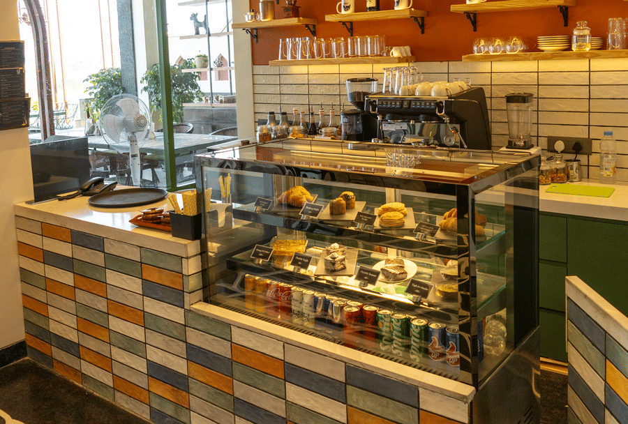 Cafe ordering section at Hotel Bawa, Udaipur showing a patterned front counter, glass food display case, coffee machine, and wall shelves.