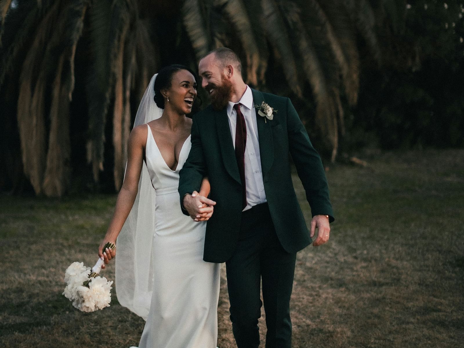 A groom and a bride walking towards the camera with a palm tree in the background