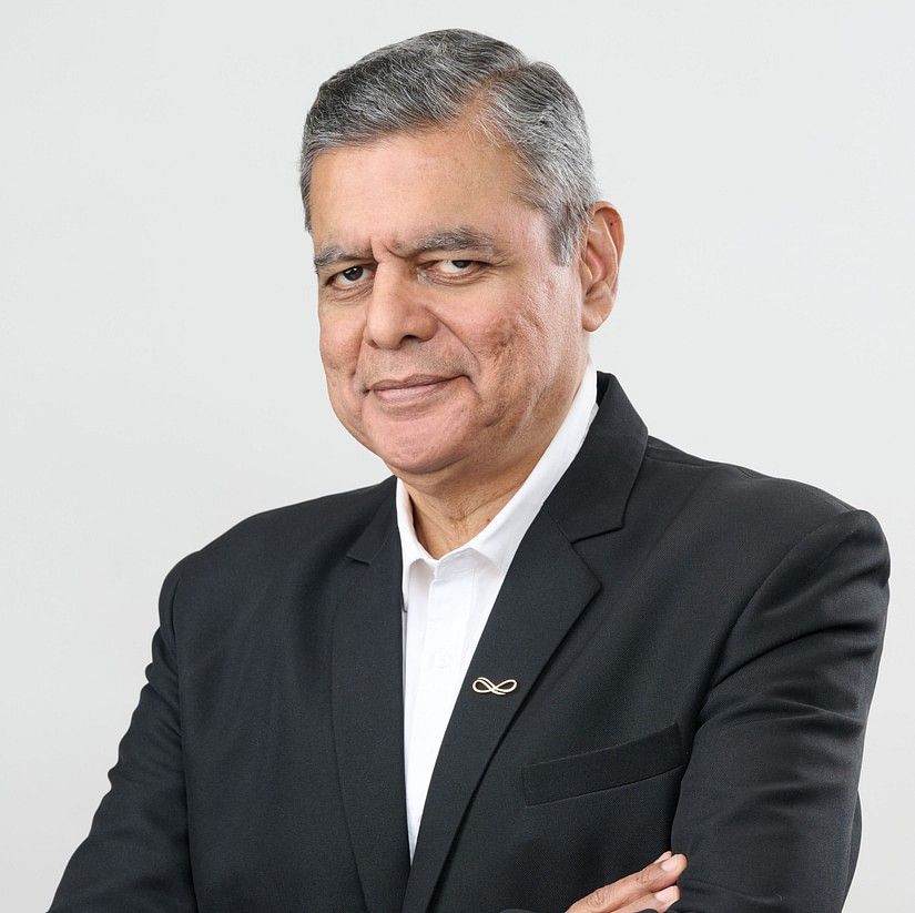 S. Raja  in a black suit and white shirt, standing with arms crossed, smiling slightly against a plain background.