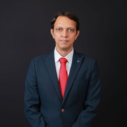 Professional portrait of a man in a dark suit with a red tie, posing formally against a black background.
