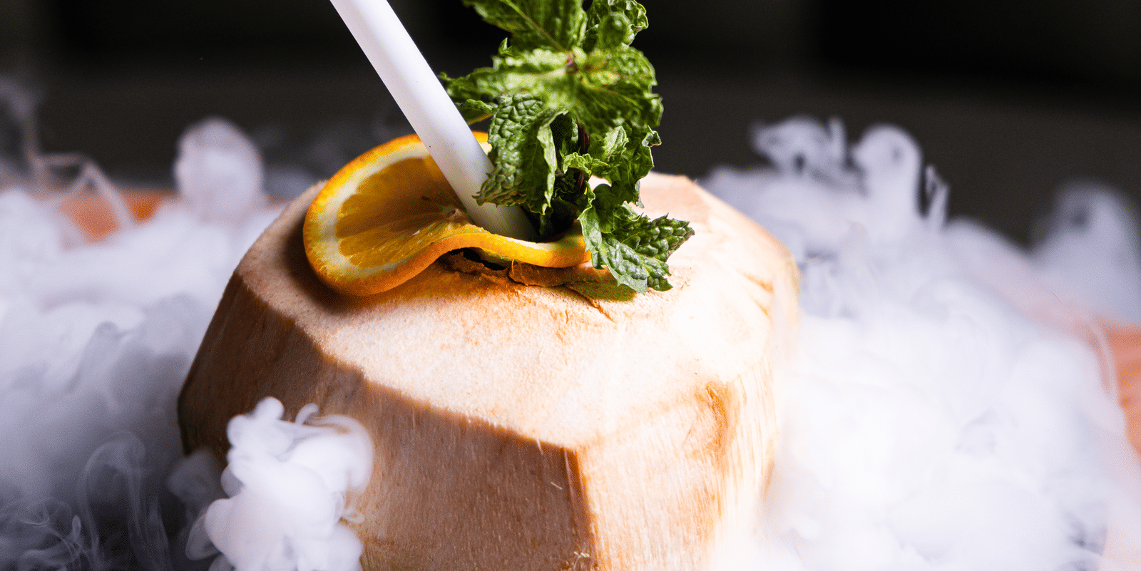 A tropical cocktail creatively presented in a coconut shell, topped with fresh mint and surrounded by smoky mist for an exotic and dramatic visual effect at Papaya Tree Hotels.