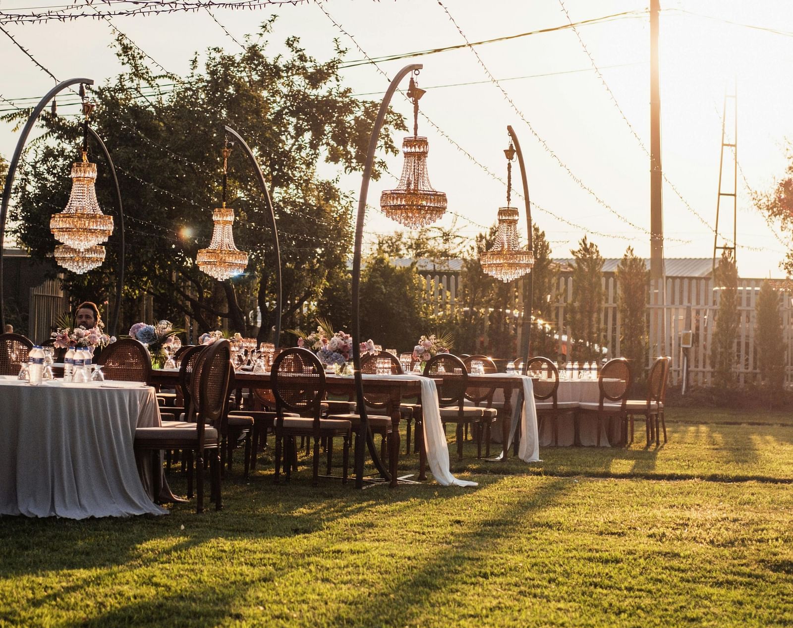 An outdoor lawn decorated with string lights, tableware and hanging lanterns at sunset.