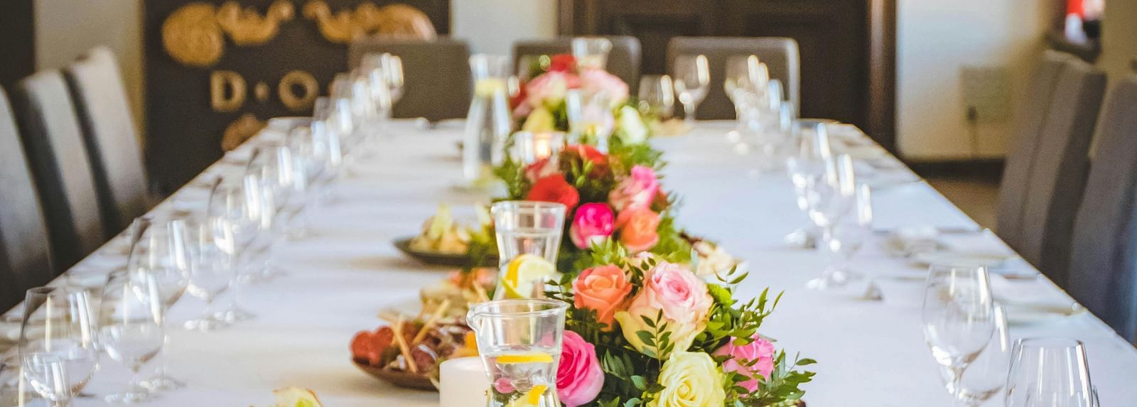 A long dining table decorated with flowers in the middle along with shiny glassware and cutlery set on the table.