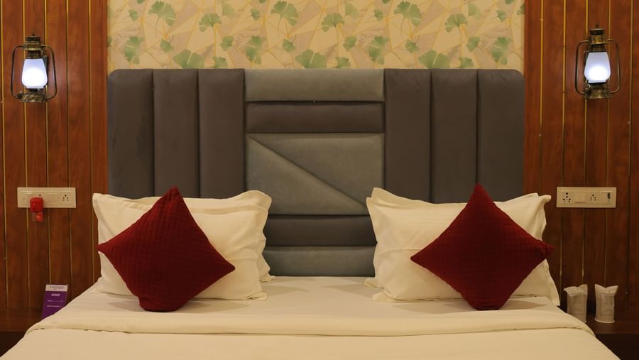 A close-up of a bed in a VITS Select Diamond Resort, Sasan Gir Executive Room. The bed has white sheets with a red bed runner and red pillows against a striped gray headboard.