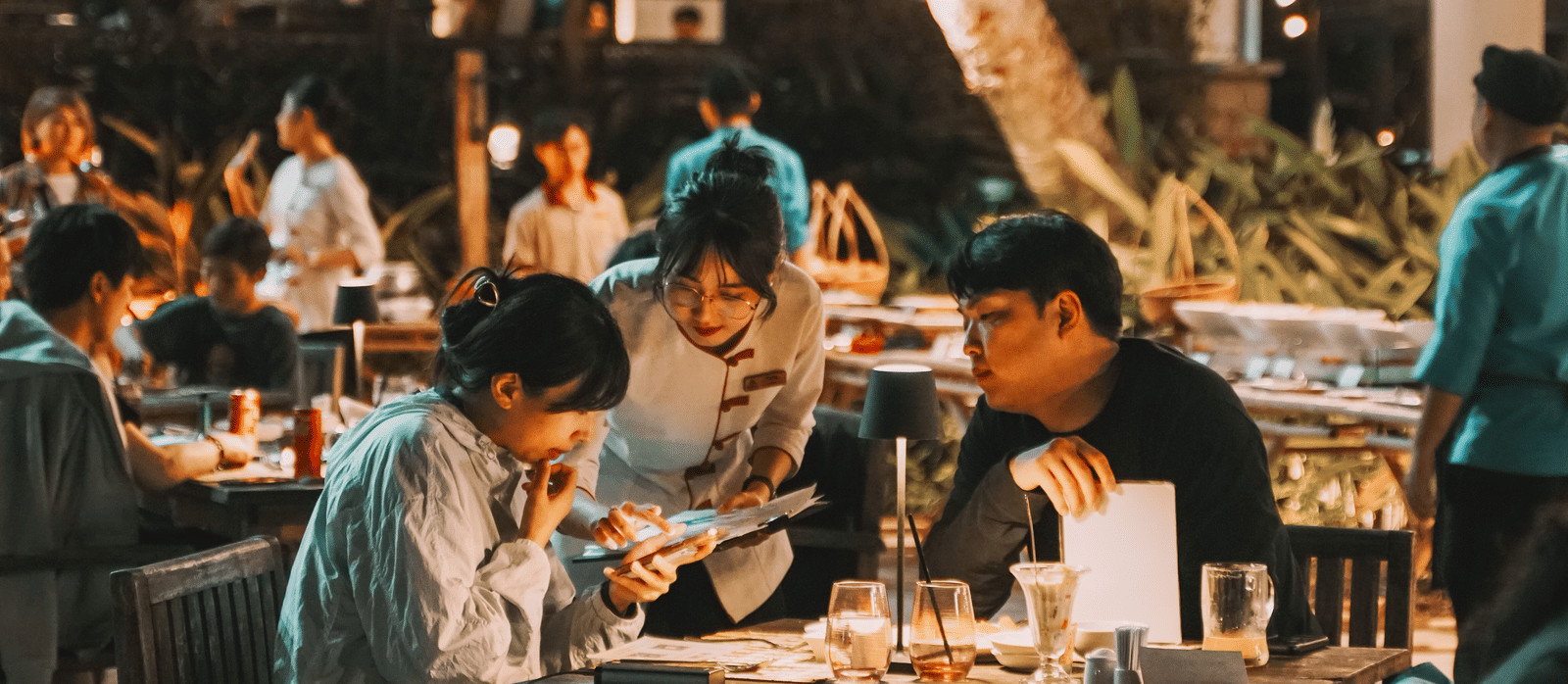 A well lit restaurant featuring people sitting on each tables and one of the staff interacting with a couple.