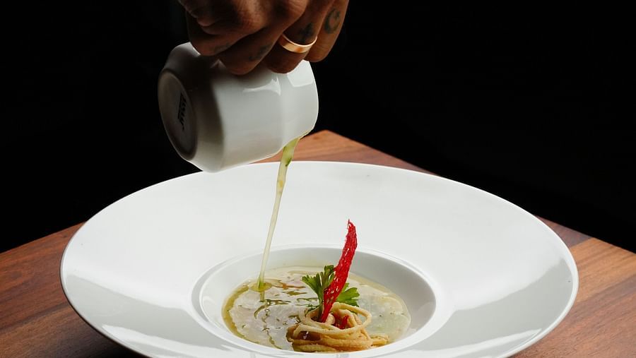 A person pouring a creamy soup into a white bowl on a wooden table at The Qualia Resort Club & Brewery, Udaipur.
