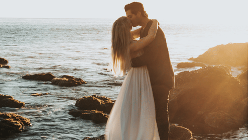 A couple embracing on top of a boulder with the ocean in the backdrop.