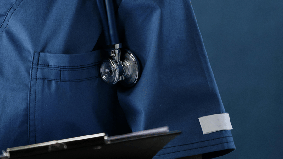 Side view of a medical worker wearing blue scrubs and a stethoscope, holding a black patient clipboard.