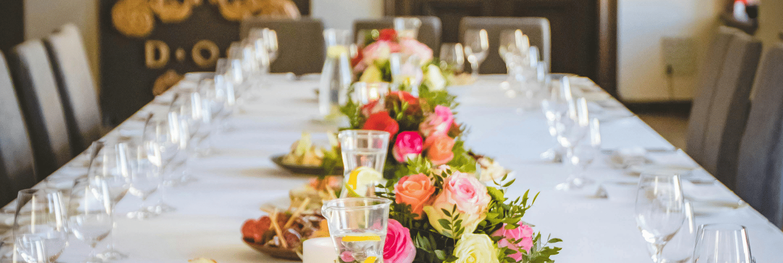 A long dining table decorated with flowers in the middle along with shiny glassware and cutlery set on the table.
