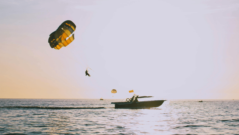 A person parasailing with a colorful parachute over calm sea waters during golden hour with a motorboat visible on the water surface and a soft pastel sky in the background.