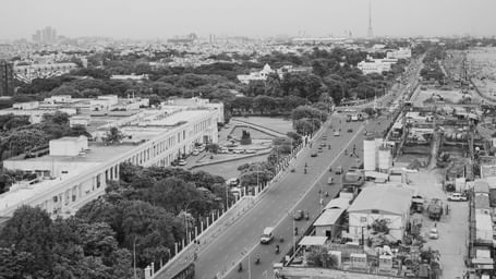 A black and white aerial photograph of Chennai shows a busy main road running through the city, flanked by residential and commercial buildings, trees, and dense urban infrastructure, with vehicles visible and the skyline extending into the distance under 