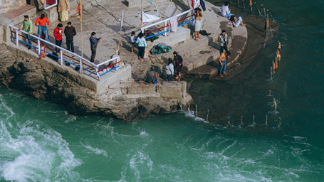 A high-angle view of a confluence, with people gathered on a stone peninsular surrounded by swirling, green waters, adjacent to a building with a pink facade.